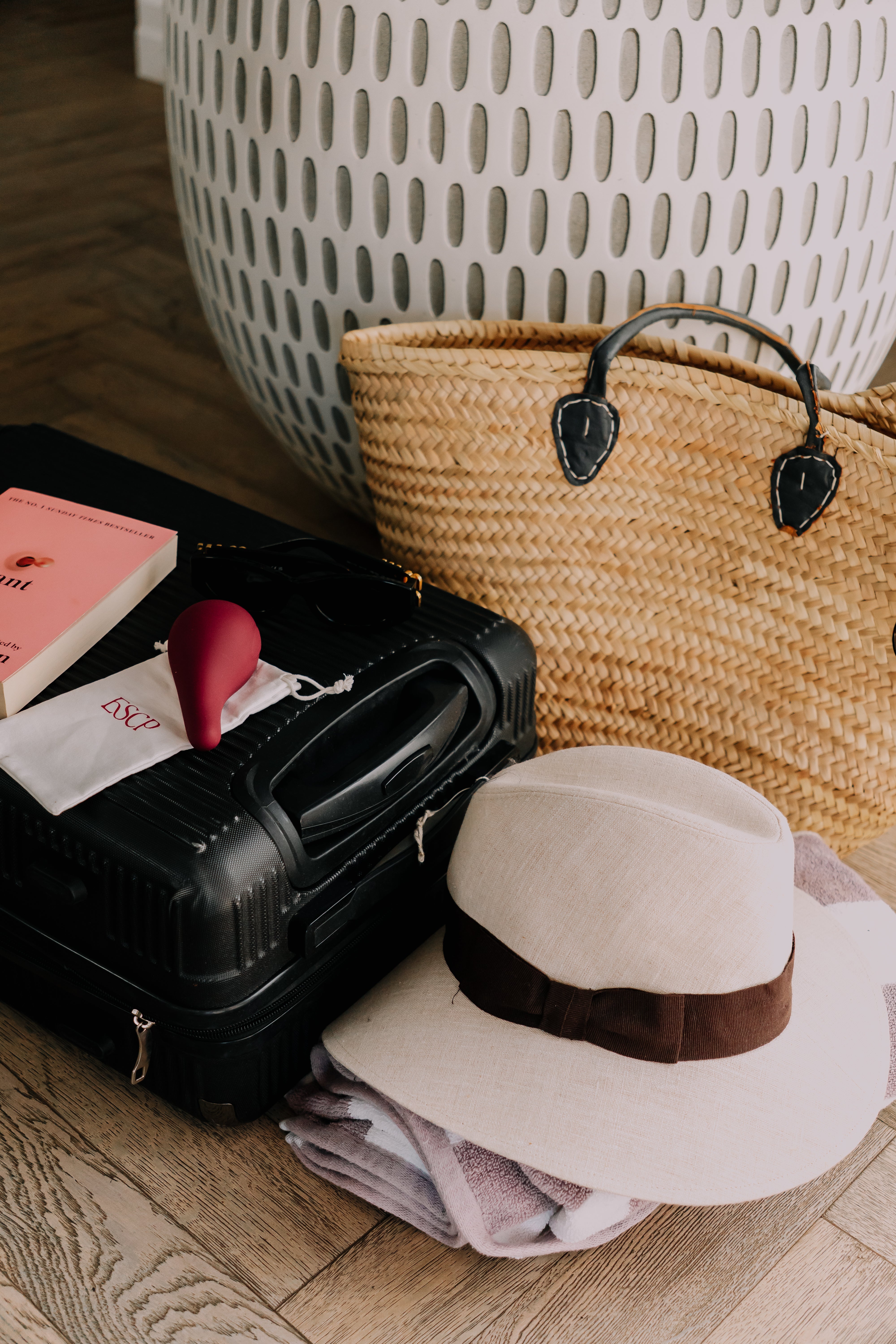 Black suitcase on a wood floor packed with a book, folded clothes, straw tote, and a cream sun hat